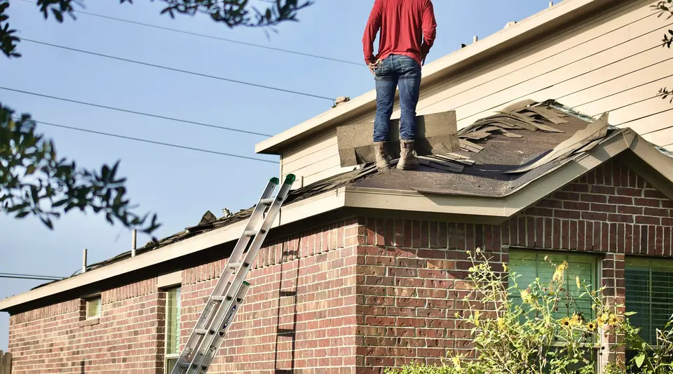 A worker standing on the roof of a brick house beside a ladder.
