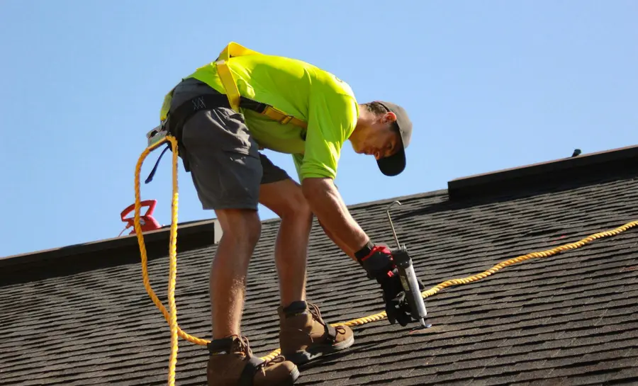 A roofer fastening shingles with a power tool and safety harness.